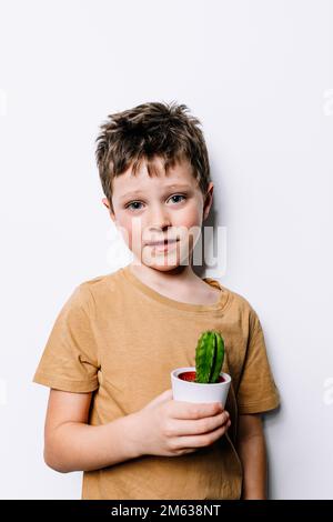 Adorabile ragazzo che tiene in pentola un piccolo cactus e guarda la fotocamera contro la parete grigia Foto Stock