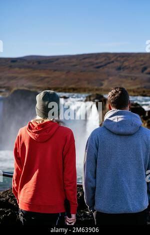 Vista posteriore di irriconoscibili viaggiatori di sesso maschile e femminile in felpe con cappuccio che si ergono sul punto panoramico e ammirano il pittoresco scenario della cascata in Islanda Foto Stock