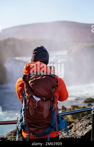 Vista posteriore dell'irriconoscibile turista maschile in cappello e felpa con cappuccio rossa in piedi sul punto di vista e scattare foto della maestosa cascata su smartphone Foto Stock