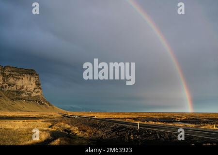 Scenario mozzafiato di arcobaleno in cielo sovrastato su prato erboso secco e strada asfaltata che conduce alla montagna rocciosa Lomagnudur in terreno vulcanico in IC Foto Stock