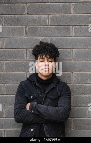 Giovane maschio latino-americano con capelli ricci in giacca nera appoggiata con braccia incrociate sul muro di mattoni ad occhi chiusi Foto Stock