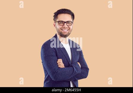 Uomo brunetto sorridente in giacca blu e occhiali con mani incrociate sul petto su sfondo beige. Foto Stock