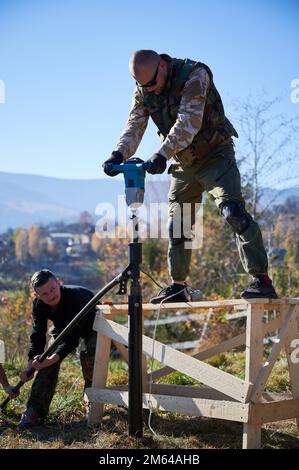 Lavoratori maschi costruzione palo fondazione per legno telaio casa. Uomini costruttori che perforano pali nel terreno su sfondo cielo blu. Foto Stock