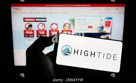 Persona che tiene il cellulare con il logo della società canadese di cannabis High Tide Inc. Sullo schermo di fronte alla pagina web aziendale. Messa a fuoco sul display del telefono. Foto Stock