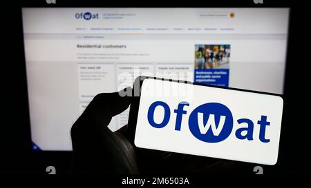 Persona che tiene il cellulare con il logo dell'autorità di regolamentazione dei servizi idrici (Ofwat) sullo schermo di fronte alla pagina web. Messa a fuoco sul display del telefono. Foto Stock