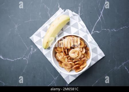 patatine di banana asciutte in una ciotola su fondo di mattonelle nere Foto Stock