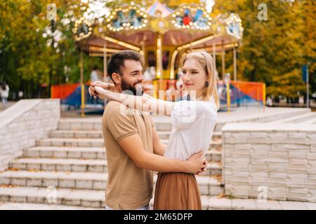 Allegra giovane coppia innamorata sorridente guardando lontano nel parco divertimenti sullo sfondo del carosello il giorno d'estate. Foto Stock