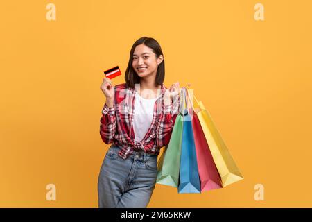 Concetto di rimborso. Donna asiatica felice posa con carta di credito e borse shopping colorate su sfondo giallo studio Foto Stock