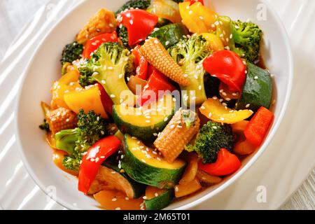 primo piano di verdure saltate in padella di peperone, cipolla, zucchine, granoturco in pannocchie, fiori di broccoli versati con salsa di soia appiccicosa in ciotola bianca su whit Foto Stock