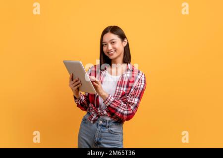 Felice donna giapponese che usa il tablet digitale e sorridente alla fotocamera, navigando in internet mentre si trova su sfondo giallo Foto Stock