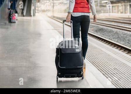 dettaglio più basso di una donna passeggero con la schiena girata portando una valigia su ruote e camminando accanto ai binari del treno alla stazione. Foto Stock