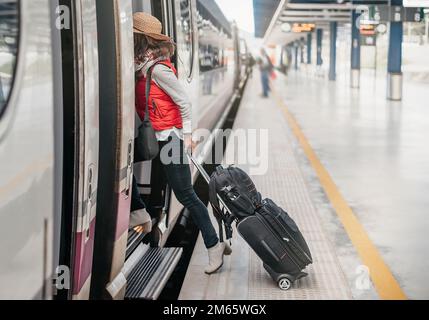 vista laterale di un turista con bagaglio a mano, entrando in un treno in attesa alla stazione. cappello e gilet rosso. viaggio in treno concept weekend. treno ad alta velocità. Foto Stock