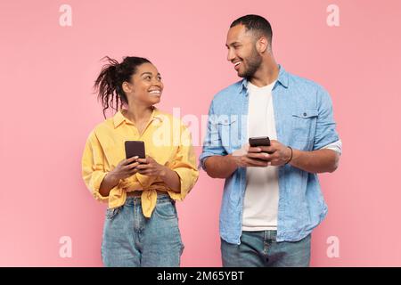 Allegra coppia nera che tiene gli smartphone e si guarda l'un l'altro, in piedi insieme su sfondo rosa Foto Stock
