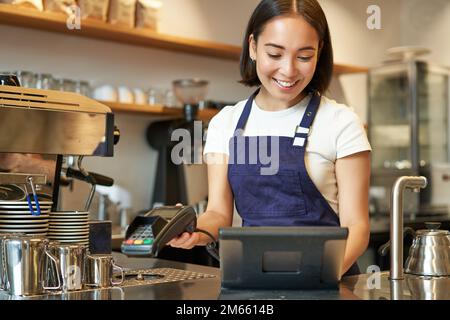 Ragazza barista asiatica sorridente che elabora l'ordine, immetti l'ordine nel terminale POS, lavora al banco del bar Foto Stock