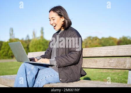 Ritratto di sorridente ragazza asiatica siede sulla panchina nel parco, parla con un amico online via laptop, video chat, utilizzando il computer per studiare in remoto Foto Stock