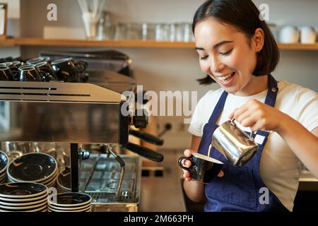 Primo piano di carina barista asiatica ragazza fare cappuccino, fare latte art in tazza con latte al vapore, in piedi in caffetteria dietro il bancone Foto Stock