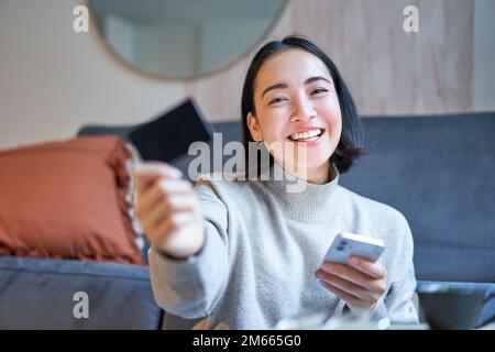 Sorridente ragazza asiatica con smartphone e carta di credito, fa il suo shopping online, utilizza il telefono cellulare per ordinare la consegna a casa Foto Stock