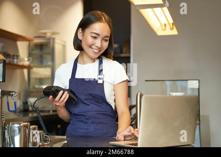 Barista ragazza asiatica sorridente, che lavora in caffè, servendo il cliente, guardando il portatile mentre si utilizza il lettore di schede per ricevere denaro per l'ordine, vendere il caffè Foto Stock