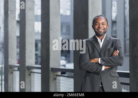Ritratto di un uomo d'affari afro-americano di successo, uomo in tuta d'affari sorridente e guardando la fotocamera da fuori ufficio edificio, boss con braccia incrociate ridere. Foto Stock