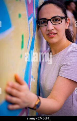 Giovane e bella donna si appoggia contro un muro colorato e si pone per la macchina fotografica - stile di strada tipico Foto Stock