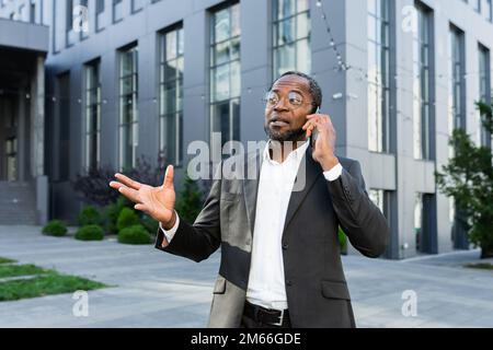 Allegro e soddisfatto boss afro-americano fuori ufficio edificio sorridendo e parlando con i colleghi al telefono, uomo in tuta d'affari a piedi durante la pausa pranzo. Foto Stock