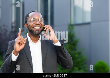 Allegro e soddisfatto boss afro-americano fuori ufficio edificio sorridendo e parlando con i colleghi al telefono, uomo in tuta d'affari a piedi durante la pausa pranzo. Foto Stock