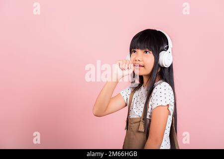 Bambino asiatico di 10 anni sorridere ascoltare musica indossare cuffie wireless e tenere la mano vicino alla bocca canta canzone Foto Stock