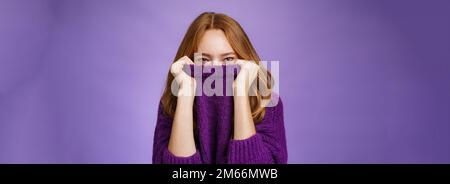 La ragazza attende l'inverno con il sorriso felice che è pronto a riscaldarsi con il maglione accogliente tirando il colletto sul viso e sorridendo con gli occhi a macchina fotografica in piedi positivo Foto Stock