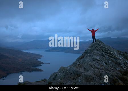 Un escursionista in giacca rossa sulla cima di una montagna si affaccia sul magnifico scenario di un lago circondato da montagne. Loch Katrine. Loch Lomond e il Parco Nazionale Trossachs. Scozia Foto Stock