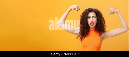 Donna sensazione potente e forte alzare le mani con pugni clenched facendo faccia intensa che si sta allenando in palestra mostrando muscoli e bicipiti guardando Foto Stock