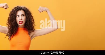 Donna sensazione potente e forte alzare le mani con pugni clenched facendo faccia intensa che si sta allenando in palestra mostrando muscoli e bicipiti guardando Foto Stock