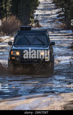 Vista frontale ad angolo del SUV fuoristrada Overland Land Cruiser che attraversa l'acqua su un sentiero innevato nella foresta Foto Stock