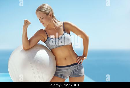 Flessibile. una giovane donna attraente flexing il suo bicep su una sfera di esercitazione dalla piscina dopo il suo workout. Foto Stock