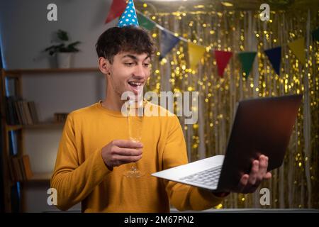 Giovane arabo che festeggia il suo compleanno con una videochiamata con gli amici usando un computer portatile. Bicchiere con champagne. Autentico Foto Stock