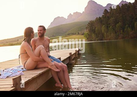 Estate idilliaca. una giovane coppia affettuosa in costume da bagno seduta sul molo al tramonto. Foto Stock