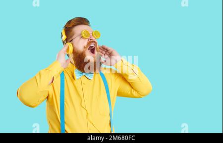 Divertente e elegante uomo grasso in cuffie ascolta musica e canta forte su sfondo azzurro. Foto Stock