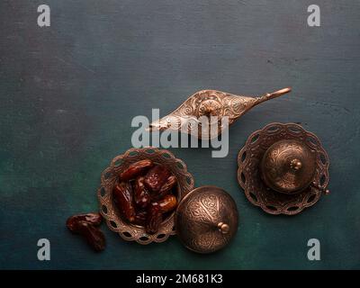 Piatto in bronzo con datteri, tazza da caffè e lampada aladdin su sfondo di legno dipinto di verde scuro con spazio vuoto per il testo. Ramadan sfondo. Giacitura piatta. Foto Stock