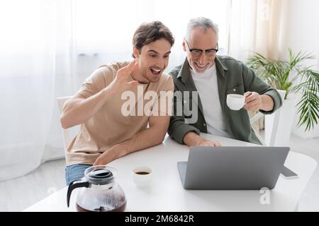 uomo eccitato che agita la mano durante la videochiamata sul portatile vicino a padre allegro con caffè tazza, immagine stock Foto Stock