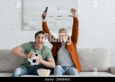 uomo maturo che mostra il gesto di vittoria e urlare vicino al figlio eccitato che tiene la palla di calcio mentre guarda la partita di calcio in tv, immagine di borsa Foto Stock