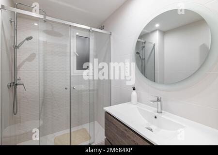 Bagno in piastrelle bianche in ceramica con grande specchio rotondo sulla parete che riflette la zona doccia con ringhiera in vetro e finiture in metallo. Foto Stock
