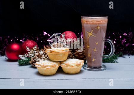 Bicchiere alto di cioccolata calda seduto accanto alle decorazioni natalizie pronto per essere appeso sull'albero con tre torte di Natale appena fatte di macinacei da avere Foto Stock