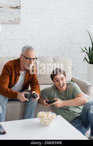 KIEV, UCRAINA - 11 MAGGIO 2022: Giovane uomo stupito gioco con papà maturo sorridente sul divano vicino popcorn Foto Stock