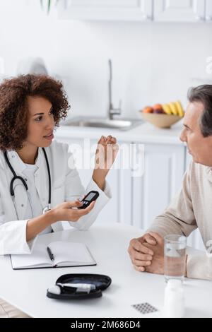curly afroamericano medico che tiene glucometro dispositivo e parlare con il paziente di mezza età, immagine stock Foto Stock