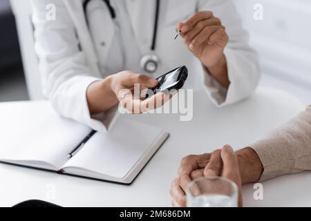 vista ritagliata del medico afroamericano che tiene il glucometro dispositivo vicino a paziente di mezza età, immagine di scorta Foto Stock