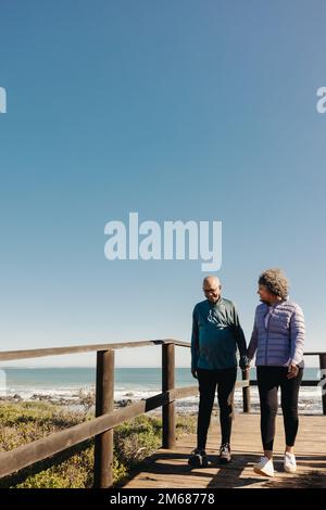Coppia anziana in pensione sorridente felicemente mentre si fa una passeggiata lungo un ponte a piedi sul mare. Coppia anziana romantica che passa un certo tempo di qualità insieme sopra Foto Stock
