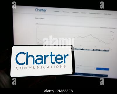 Persona che tiene il telefono cellulare con il logo della società americana di media Charter Communications Inc. Sullo schermo di fronte alla pagina web. Messa a fuoco sul display del telefono. Foto Stock