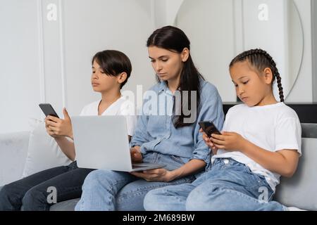 I bambini asiatici che utilizzano i cellulari mentre la madre lavora sul portatile a casa, immagine di scorta Foto Stock