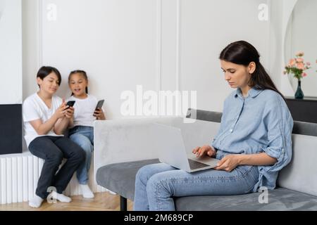 Madre che usa un notebook vicino a bambini asiatici sfocati con cellulari a casa, immagine di scorta Foto Stock