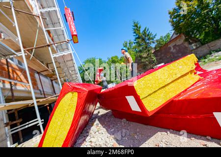 La pila di materiale termoisolante è avvolta con un foglio di cellofan. Lavoratore appende un pacchetto con lana di roccia sul gancio della gru, che lo solleva Foto Stock