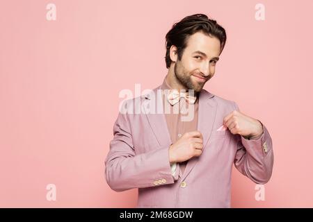Sorridente ospite di evento in giacca toccare fazzoletto isolato su rosa, immagine stock Foto Stock